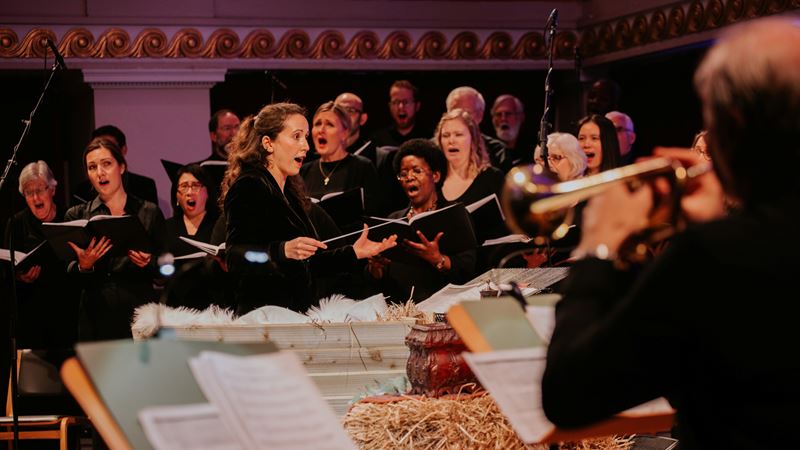 Singalong to iconic carols at All Souls Carols in the West End. Credit: Tuuli Platner. Image courtesy of All Souls. A photo of carol singers holding books with lyrics while singing together and a photo of a nativity set in the foreground with a person playing a brass instrument