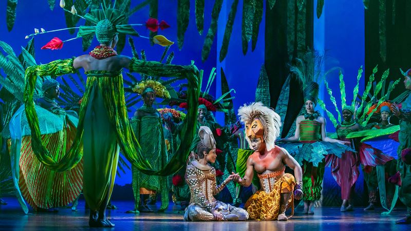 Revisit one of Disney's best-loved animated films, The Lion King, with its long-running musical theatre production, currently at the Lyceum Theatre. Credit: Disney. Image courtesy of Disney. Actors portraying Simba and Nala hold hands while other members of The Lion King musical cast surround them, dressed and posing as luxurious and tropical trees and plants.