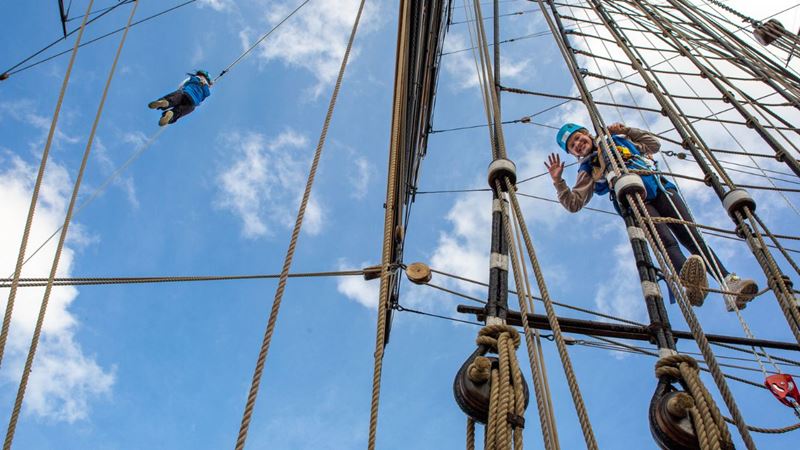 Experience panoramic views across London's skyline at the Cutty Sark Rig Climb Experience. Image courtesy of Royal Museums Greenwich. Photo credit: Nathan Turner. a low angle shot of people wearing jumpsuits and harnesses, climbing the Cutty Sark with a blue sky in the background