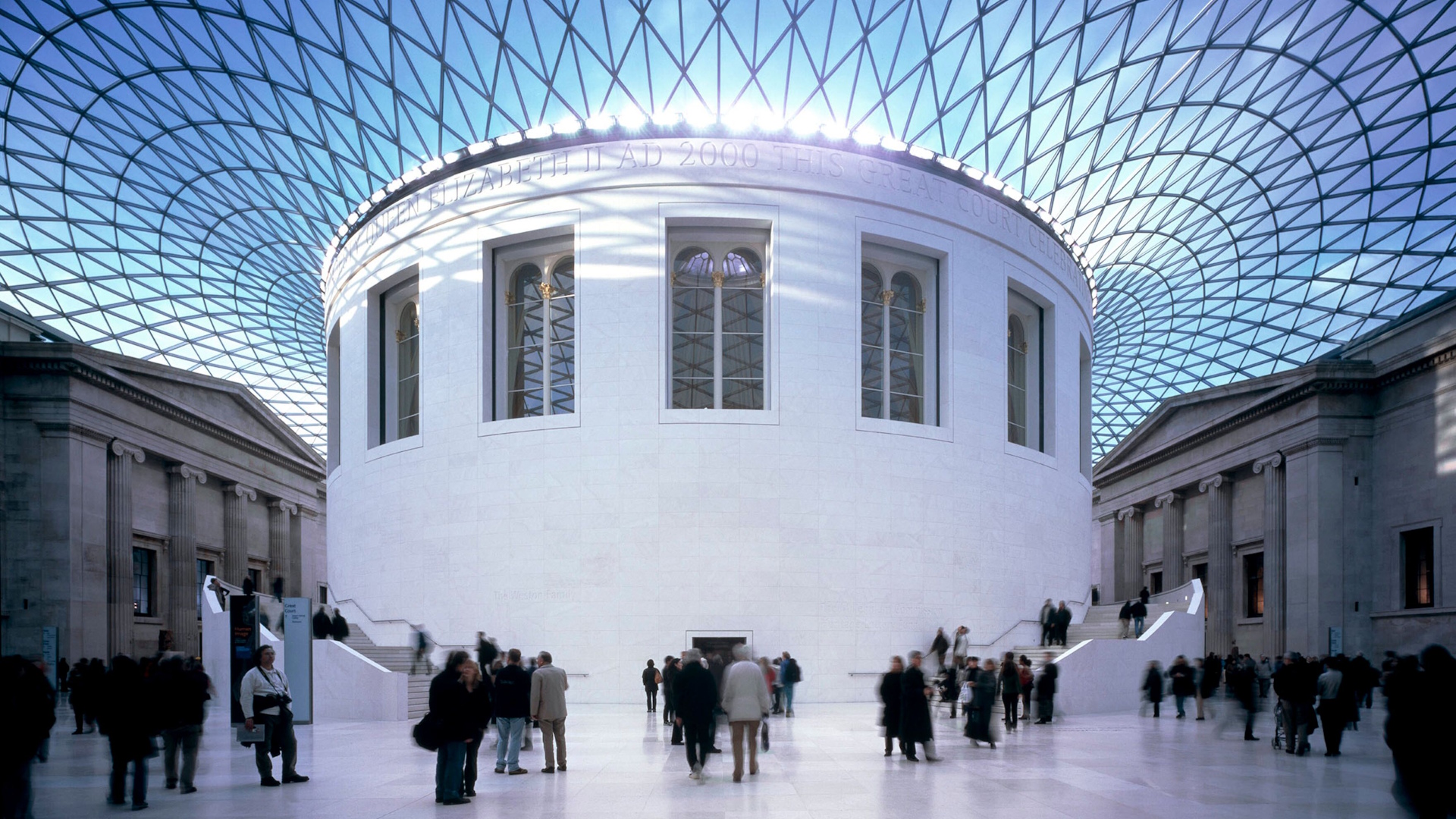 The British Museum. Credit: Trustees of the British Museum. Image courtesy of the British Museum. The Great Court at the British Museum with blue sky and light peering through the glass rood, round white structure in the middle, visitors all around and Greek-style buildings in the background.