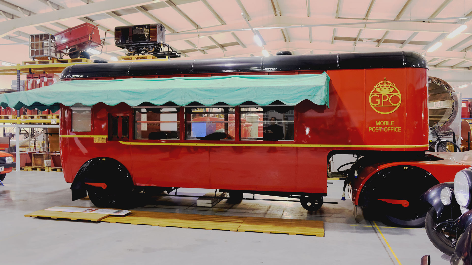 Immerse into the rich history of the Post Office by viewing rarely exhibited vehicles and equipment at the Postal Museum Store in London. Photo credit: Carole Rocton A large red mobile Post Office trailer with a green cover, acting as a post office complete with a phone booth and postal services, is on display at the Postal Museum Store.