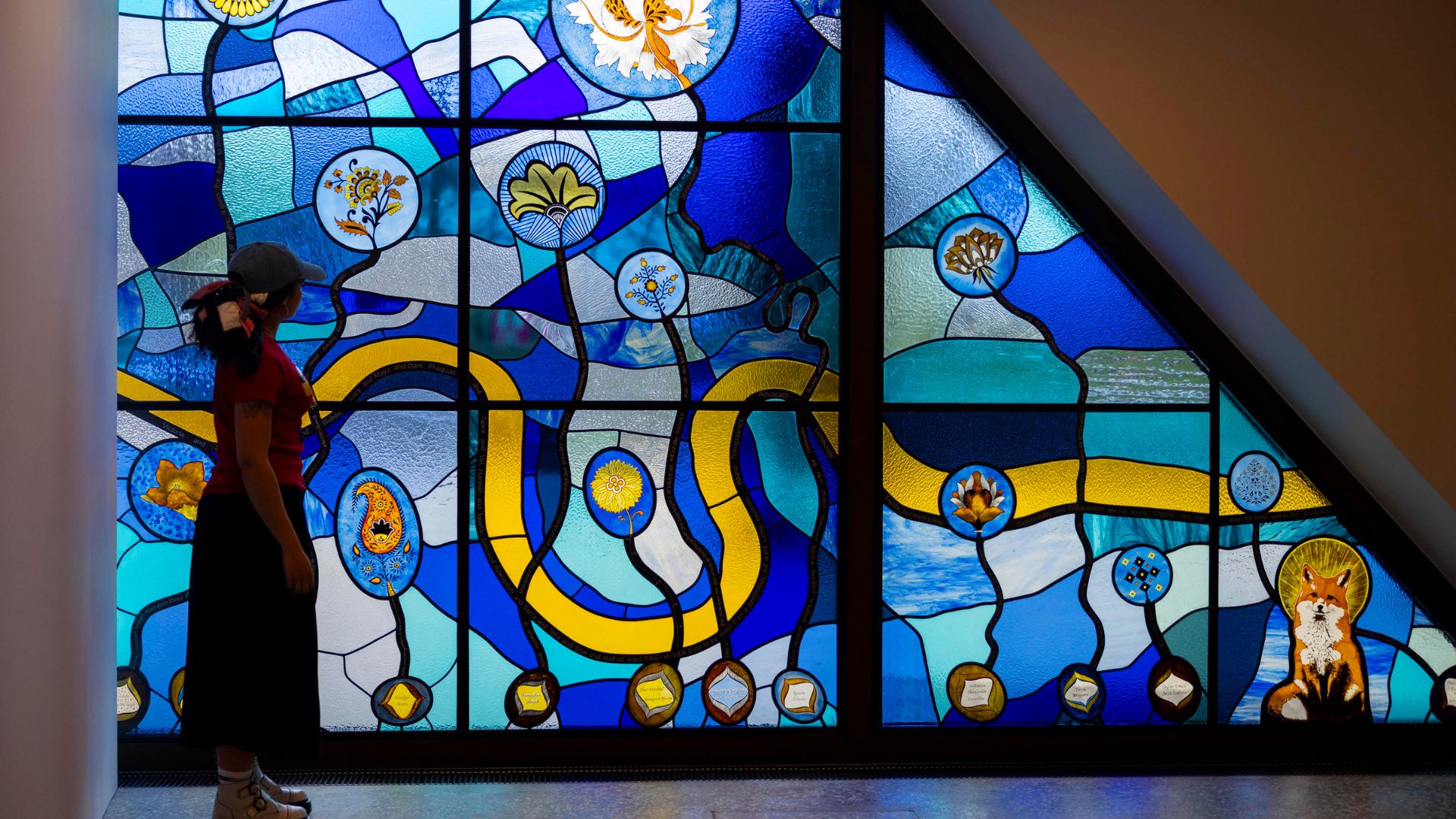 Discover the New Work Commission by Tania Bruguera for V&A East Museum © David Parry for the V&A. Image courtesy of V&A East. Blue patterned stain glass window with person looking out to it.