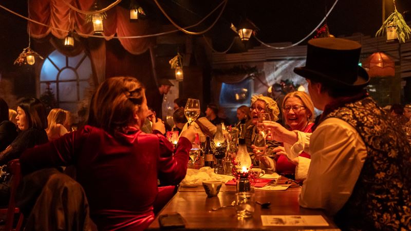 Enjoy food, theatre and music with a festive twist during The Great Christmas Feast. Credit: The Lost Estate. Image courtesy of The Lost Estate. A group of guests dressed in festive themed clothing sat at a table raising their glasses in a festive themed room surrounded by actors
