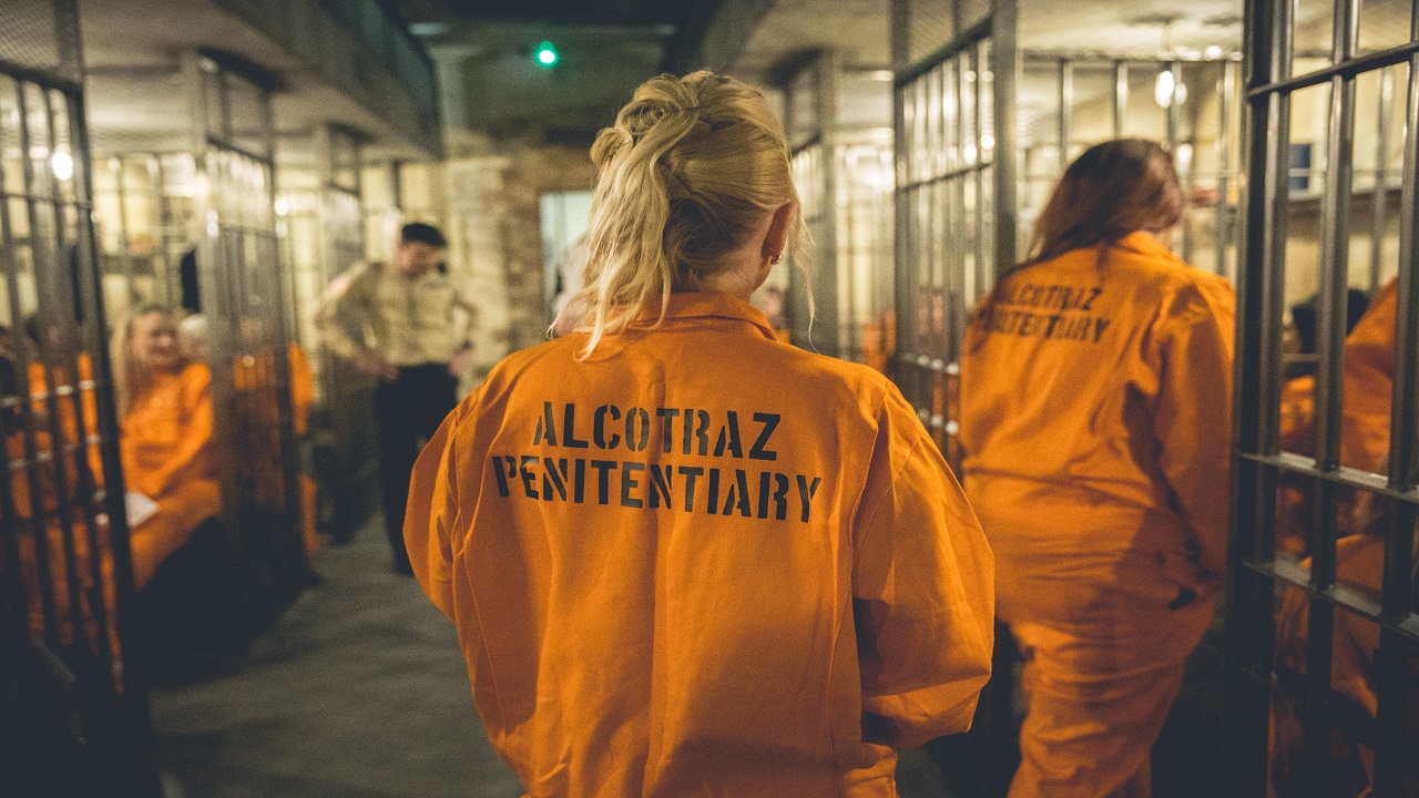Experience the unique immersive experience at Alcotraz Prison Cocktail Experience in London. Image courtesy of Golden Tours. a woman wearing a orange prison jumpsuit with 'Alcotraz Penitentiary' printed on the back, walking through the different prison-themed cells