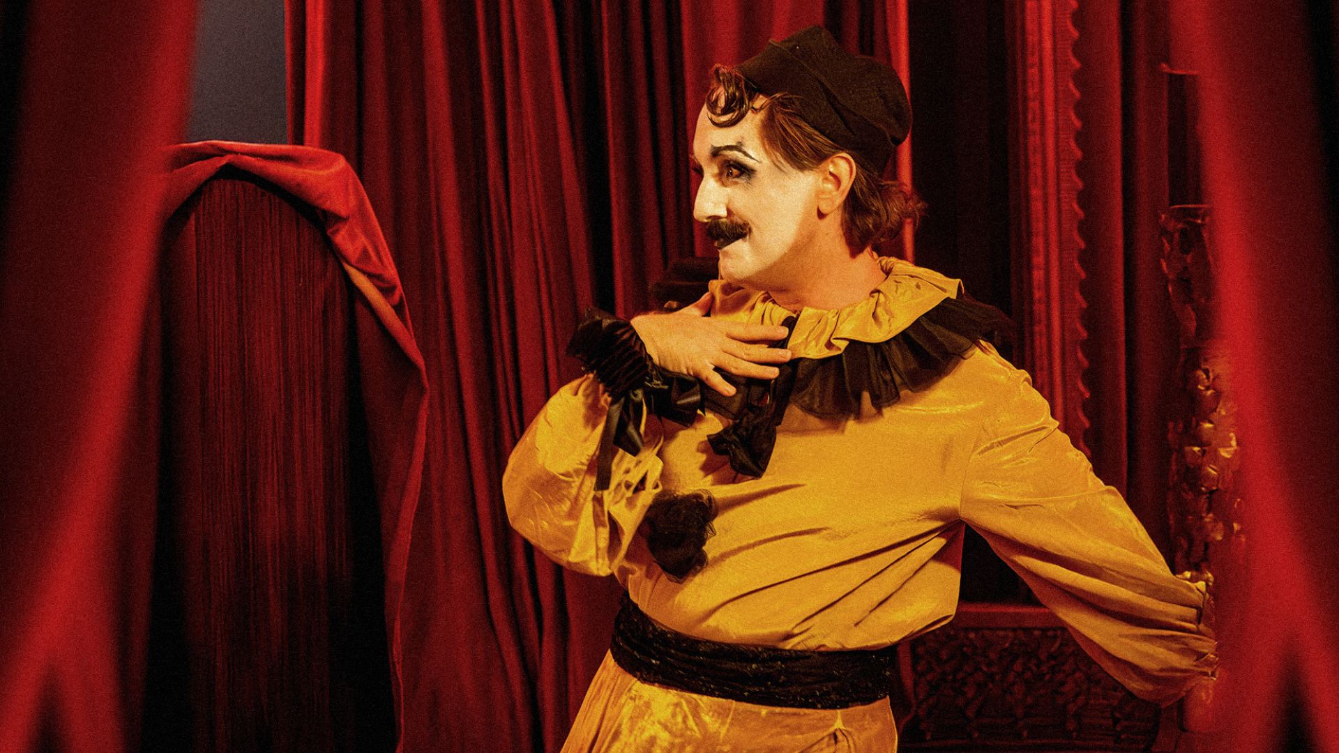 The stage is set for drama and delight – discover the bold characters that bring CHAT NOIR to life. Image courtesy of The Lost Estate. A performer in a striking yellow costume with black accents posing dramatically against deep red velvet curtains at CHAT NOIR