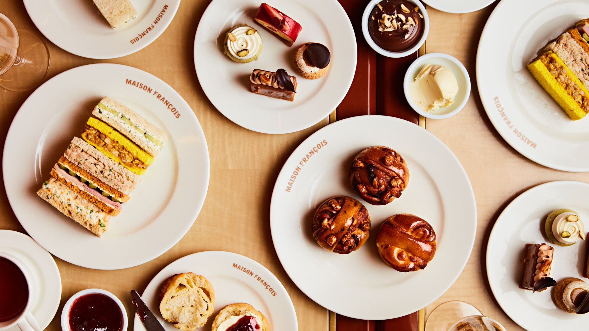 Enjoy a French twist on a traditional afternoon tea at Maison François. © Steven Joyce/Maison François Overhead view of a Maison François afternoon tea featuring finger sandwiches, babkas with jam and cream, pastries and chocolate desserts served on branded white plates.