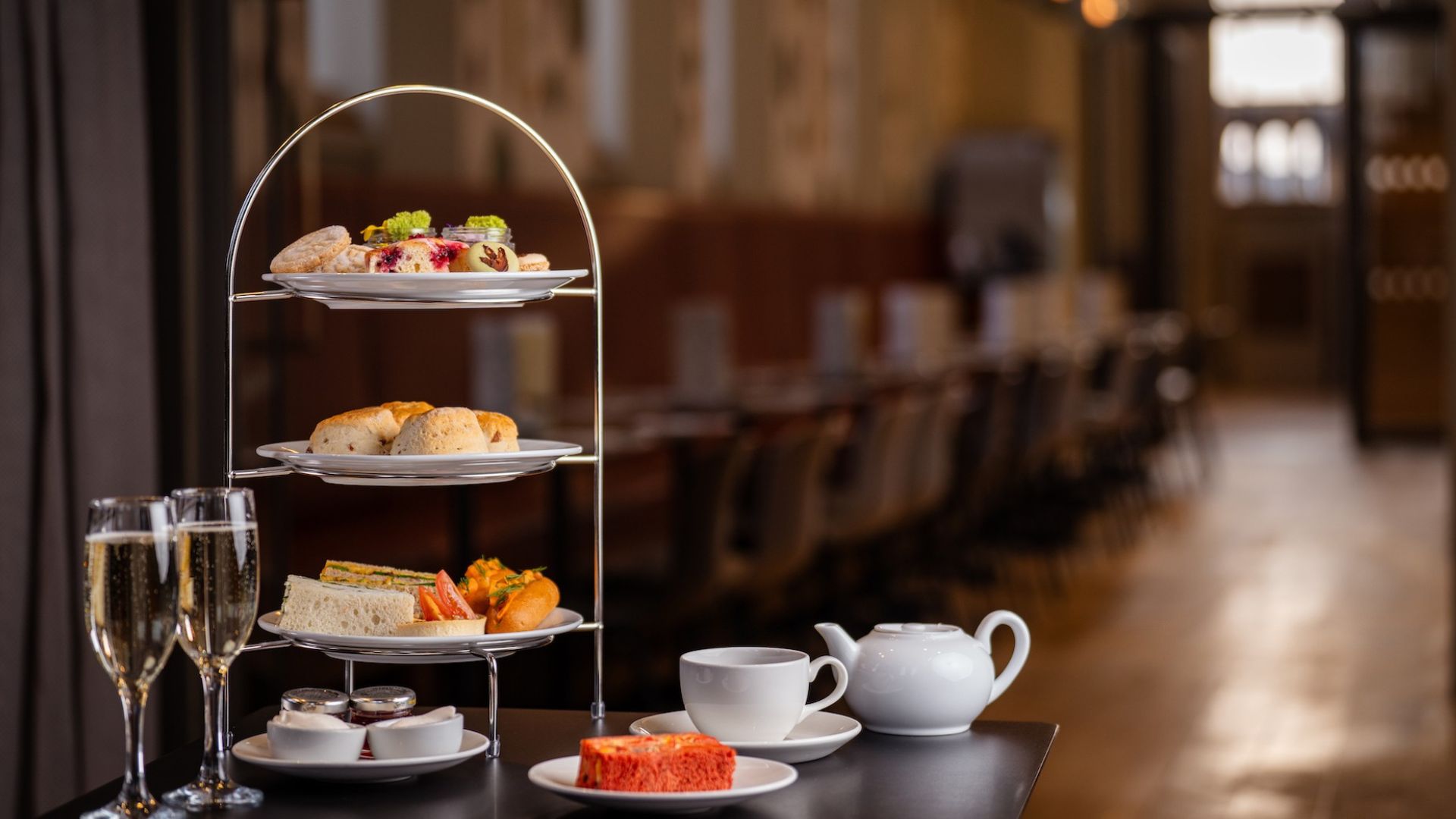Tuck into an elegant afternoon tea at the Natural History Museum with sandwiches, scones and themed cakes and sweet treats, served in a stylish dining room. Image courtesy of Benugo. Three-tier afternoon tea stand with finger sandwiches, scones and cakes on a table alongside a teapot, teacup and two glasses of Champagne in a restaurant setting.