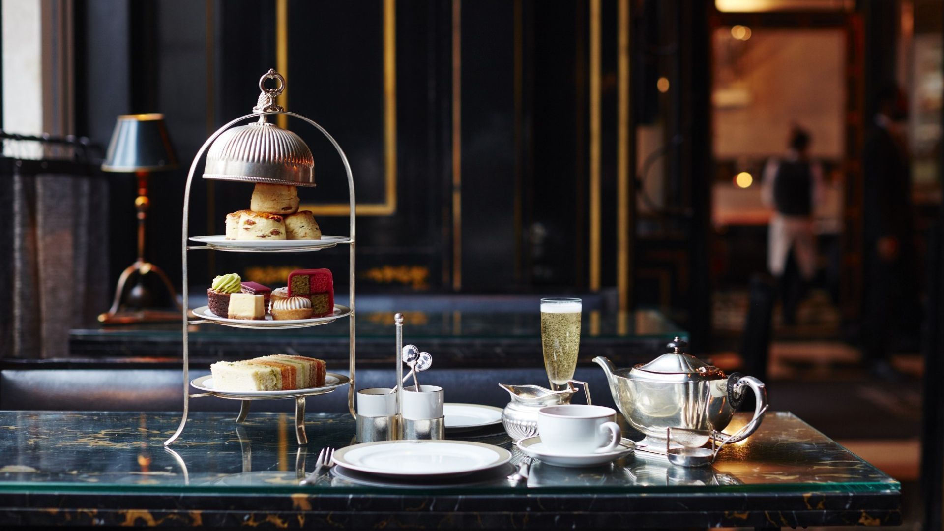 Enjoy an indulgent afternoon tea, featuring delicate sandwiches, freshly baked scones and elegant pastries, at The Wolseley. Image courtesy of The Wolseley. Three-tier afternoon tea stand with scones, cakes, and finger sandwiches on a marble table, alongside a silver teapot, teacup, and a glass of sparkling wine in an elegant, softly lit dining room.