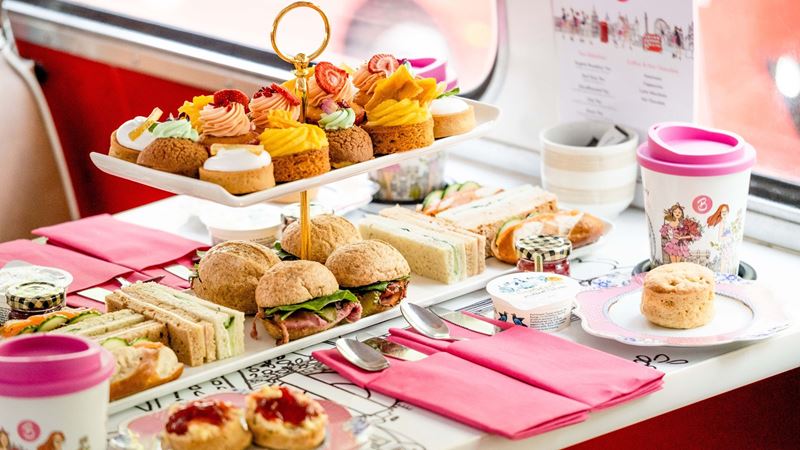 Tuck into sweet treats and savoury delights and spot famous London landmarks onboard Brigit's Bakery afternoon tea bus tour. Image courtesy of Brigit's Bakery. A table on a double decker bus covered in sandwiches and cakes with coffee cups.