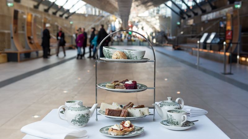 Enjoy a delicious afternoon tea beneath the iconic copper-clad hull of the Cutty Sark. Credit: Red Letter Days. Image courtesy of Red Letter Days. an afternoon tea stand on a table with small plates of sandwiches and scones, and teapots and saucers either side, set in a museum space