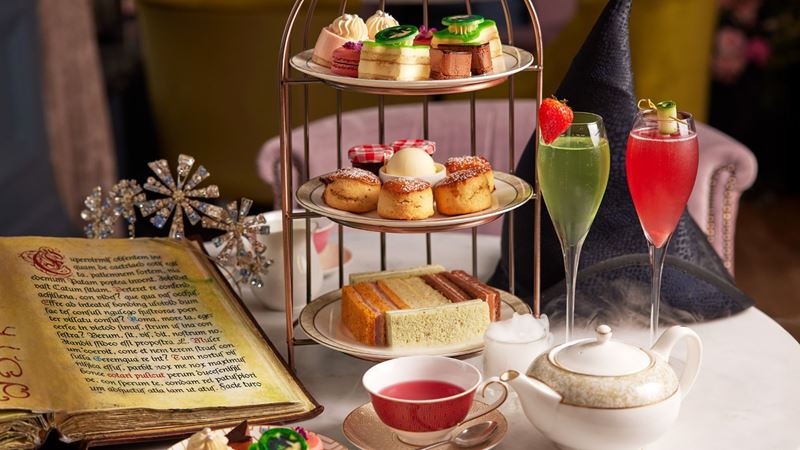 A themed Wicked afternoon tea served at The Clermont London, Victoria, featuring colourful cakes, sandwiches and theatrical cocktails inspired by the musical. Image courtesy of The Clermont Hotel. A tiered afternoon tea stand with cakes, scones and sandwiches beside colourful cocktails and a teapot on a decorated table.