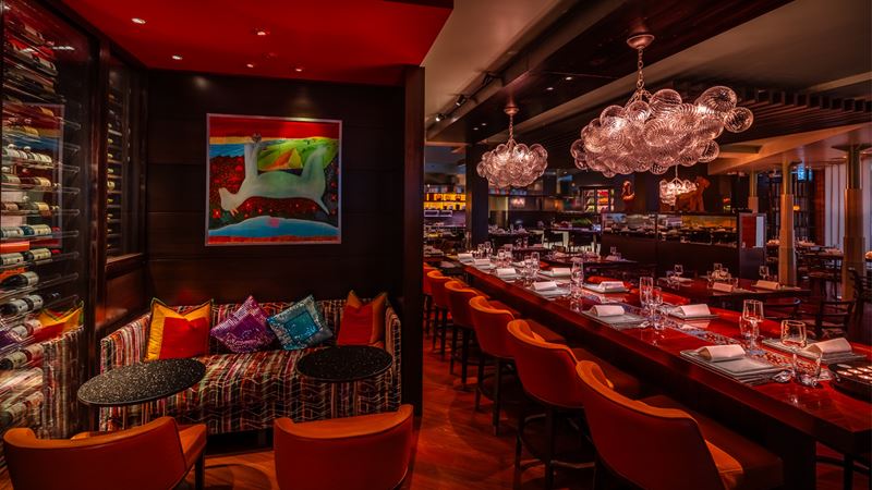 See the chefs at work from the open kitchen at Michelin-star Amaya. Credit: Amaya. Image courtesy of Amaya. Darkly lit Amaya restaurant interior, featuring red carpeting, brown walls, a lounge area with colourful sofa, red seats and cushions, and a counter with red bar stools, plates and glasses.