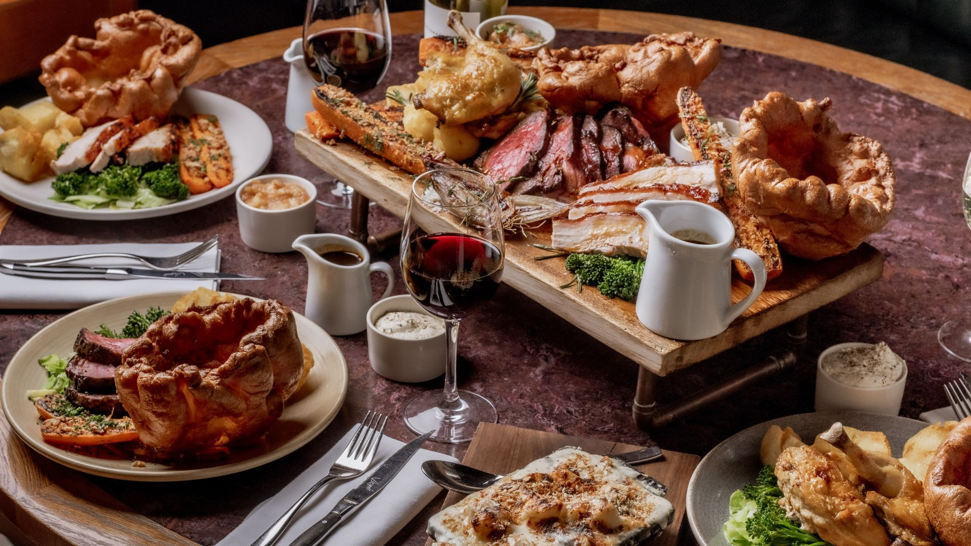 A classic Sunday roast becomes a true feast at Bread Street Kitchen & Bar, where towering Yorkshire puddings, golden roast potatoes and generous cuts of meat make family lunches something to savour. Image couresty of Gordon Ramsay. A large Sunday roast spread with Yorkshire puddings, roasted meats, vegetables and gravy served on a table.