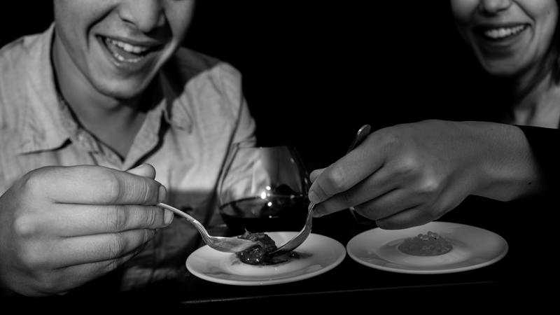 Ignite the senses during a meal in total darkness at Dans Le Noir. Credit: Romain Ruiz. Image courtesy of Dans Le Noir. A black and white photo of a man and woman scooping up food to eat in darkness with smiles on their faces