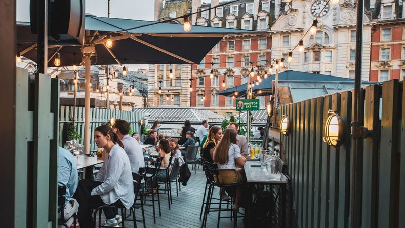 Lively food halls such as Market Halls bring the buzz of London dining to families, with rooftop terraces, long tables and a choice of street-food favourites for every taste. Image courtesy of Market Halls. People sitting at tables on a rooftop terrace with string lights and umbrellas at a busy food hall.