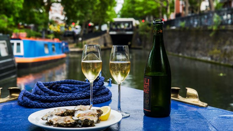 Canal cruise with a view. Credit: Haarala Hamilton Photography. Image courtesy of London Shell Company. A landscape view of a seafood platter and bottle of wine on a barge, with the canal in the background blurred.