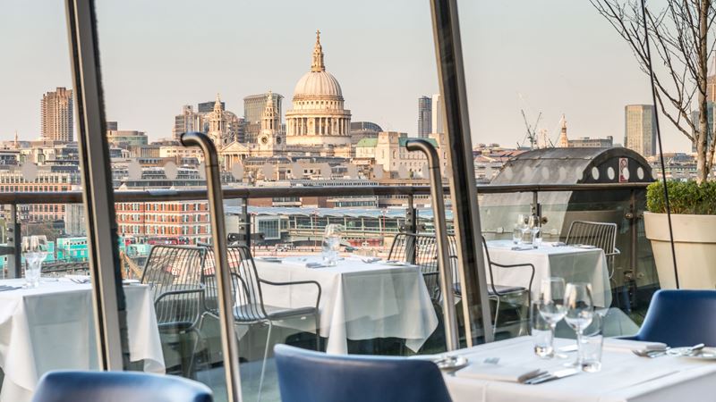 Take in the sweeping views of St Paul's Cathedral from OXO Tower Restaurant. Credit: Oxo Tower Restaurant, Bar and Brasserie. Image courtesy of OXO Tower Restaurant, Bar and Brasserie. Restaurant tables with white tablecloth, wine glasses and blue chairs overlooking the London skyline with St Paul's Cathedral in the background.