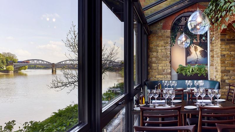 Enjoy Rick Stein’s tranquil restaurant along Barnes’ riverside. Credit: Kilian O'Sullivan. Image courtesy of Rick Stein. Rick Stein's restaurant in Barnes with wooden tables and chairs, brick walls and big open windows overlooking the river Thames.