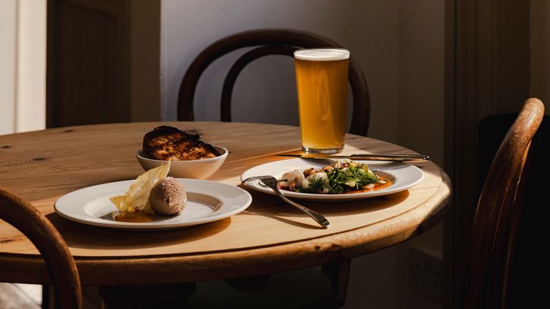 Discover London's newest Michelin-star pub, The Kerfield Arms, in Camberwell. Credit: Joe Howard. Image courtesy of The Kerfield Arms. Wooden table and chairs with a pint of beer, an elegant seafood plate, some bread in a basket and a chocolate mousse dessert.