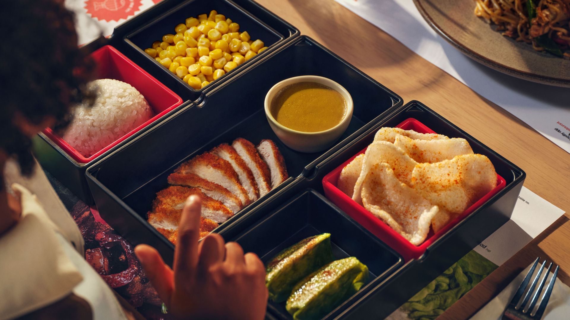 A colourful kids’ bento box served at Wagamama, featuring rice, crispy chicken, vegetables and snacks designed for younger diners. A children’s bento box meal with rice, sweetcorn, crispy chicken, dipping sauce and snacks arranged in separate compartments on a tray.
