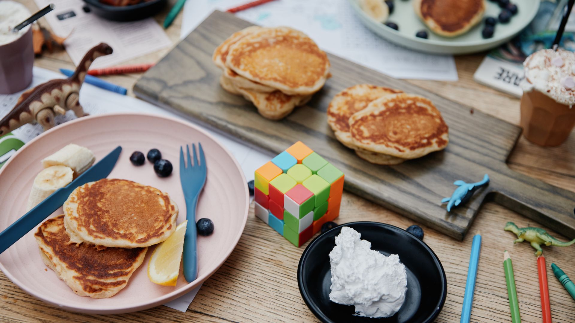 Slow mornings, messy tables, and pancakes shared between bites of laughter at Where The Pancakes Are. Image courtesy of Where The Pancakes Are. A cosy breakfast scene with stacked pancakes on plates, blueberries and banana slices, a lemon wedge, and a dollop of cream, surrounded by children’s toys, crayons, and a Rubik’s cube on a wooden table.
