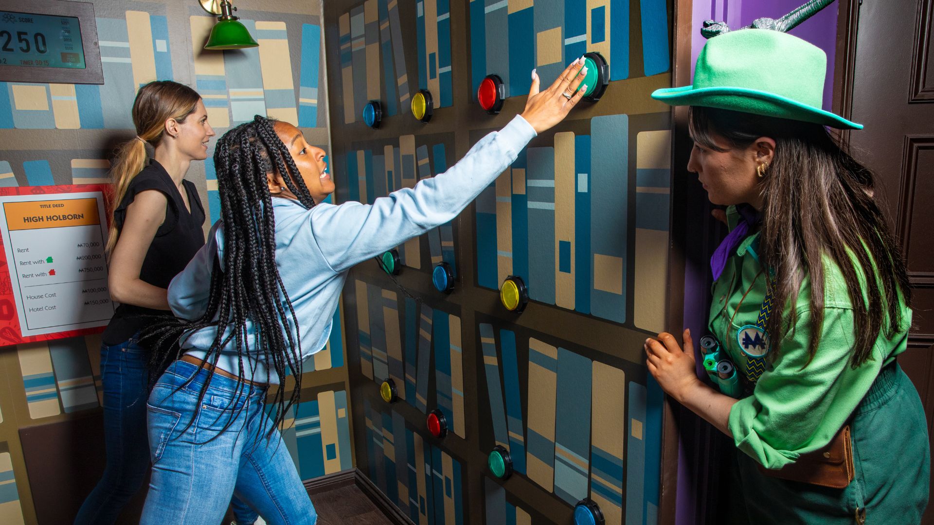 Conquer escape room challenges and dominate a life-sized board at Monopoly Lifesized. Image courtesy of Monopoly Lifesized. two women completing an escape room challenge pressing buttons on a wall, whilst a character from the experience looks on from the side