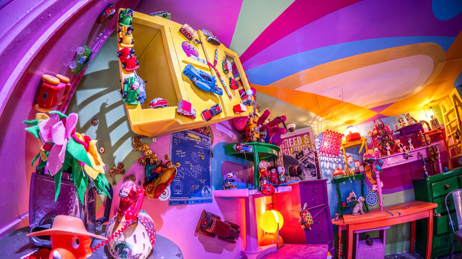 Explore a world beyond ordinary at Absurd City. Image courtesy of Wake The Tiger. A brightly coloured room inside Wake The Tiger's immersive experience, filled with toys, artwork, colourful furniture and quirky objects beneath a rainbow-striped ceiling.