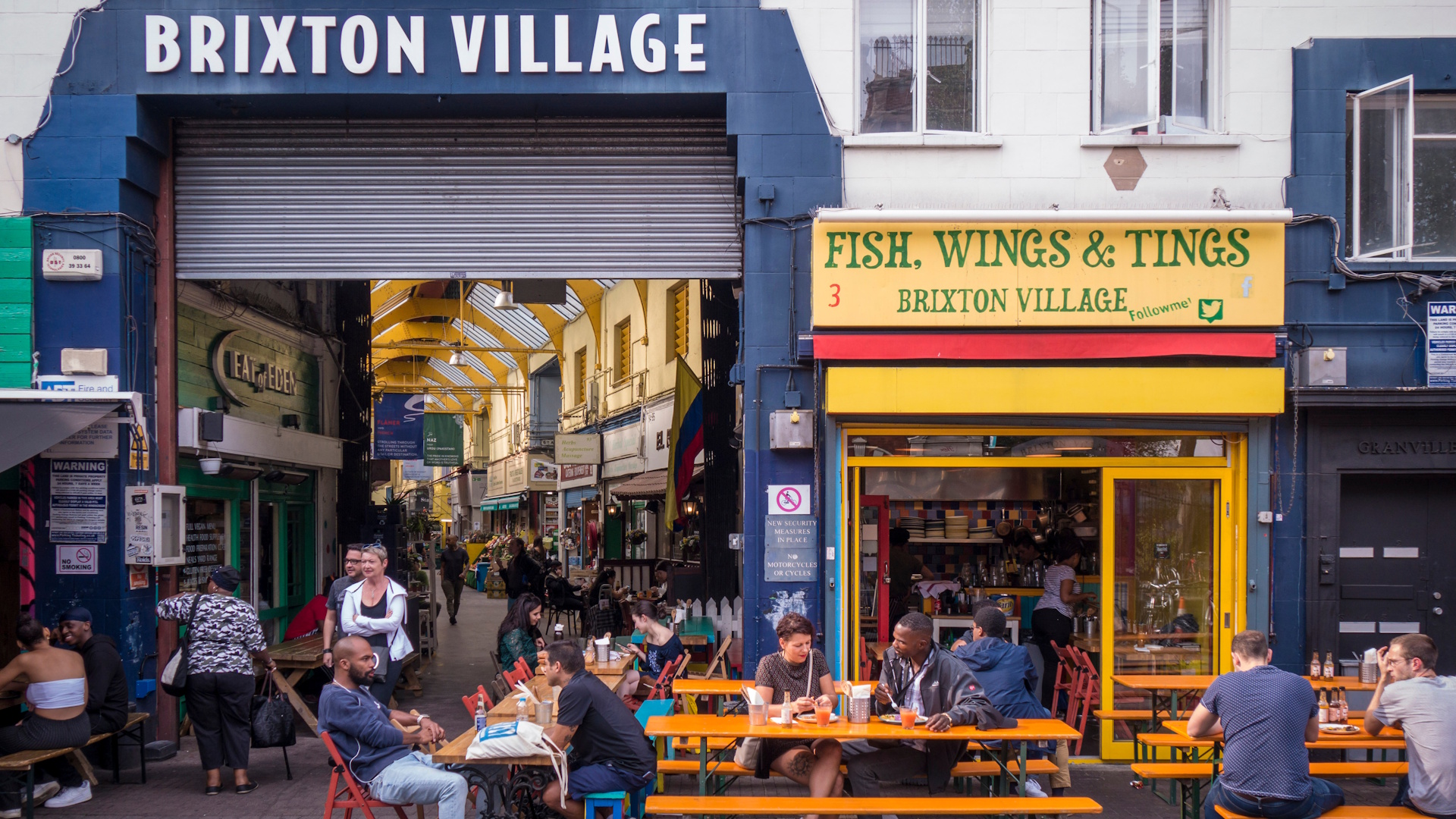 Wander into the covered arcades of Brixton Village for foodie delights galore. Credit: Shutterstock. Image courtesy of Shutterstock. Entrance framed by dark blue sign to the covered arcade of Brixton Village, with Fish, Wings and Tings yellow-fronted restaurant next to it.
