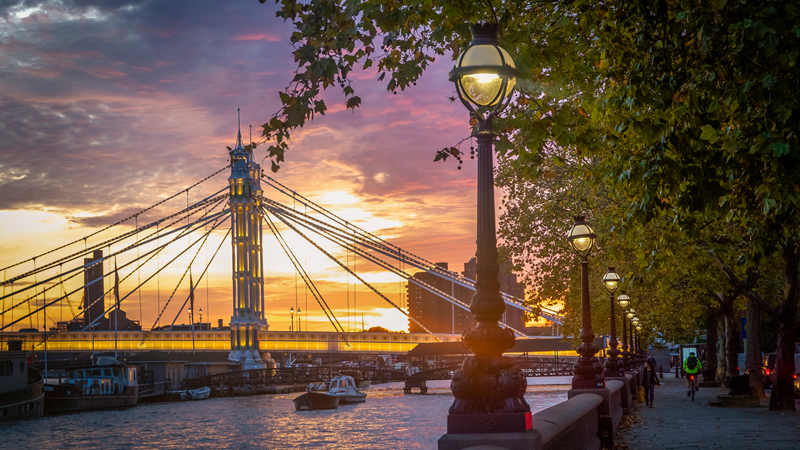 Admire the view of the Albert Bridge that runs over the river Thames. Credit: Shutterstock. Image courtesy of Shutterstock. A view of the Albert Bridge from Chelsea Embankment with a colourful sunset in the background of yellow, pick and purple with lamps and trees in the foreground