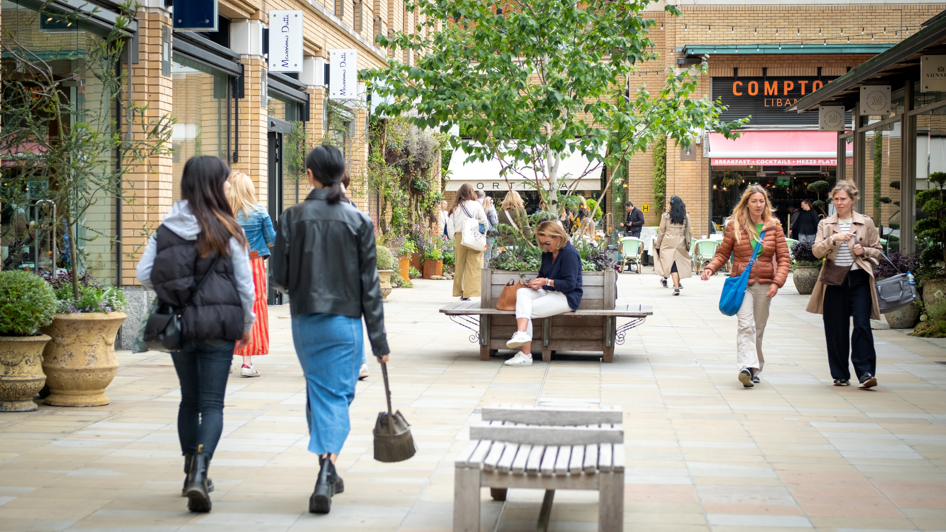 Wander around Duke of York Square in Chelsea, an ideal shopping spot. Credit: Shutterstock. Image courtesy of Shutterstock. Two women walk away from the camera at Duke of York Square with people in the distance, benches, trees and shops and restaurants in the background