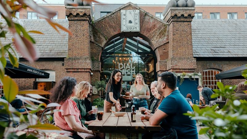 Flick through the menu of seasonal dishes, pizzas and roasts at the Dial Arch in Woolwich. Credit: Visit Greenwich. Image courtesy of Visit Greenwich. A photo of a group of friends sat on a bench at the Dial Arch pub garden as a waitress lays food down