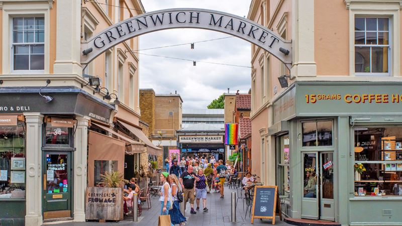Browse the stalls selling street food, vintage clothes, crafts and more at Greenwich Market. Photo by Ed Simmons. Image courtesy of Visit Greenwich. A photo of the entrance to Greenwich Market with people walking around holding shopping bags and a sign overhead that reads