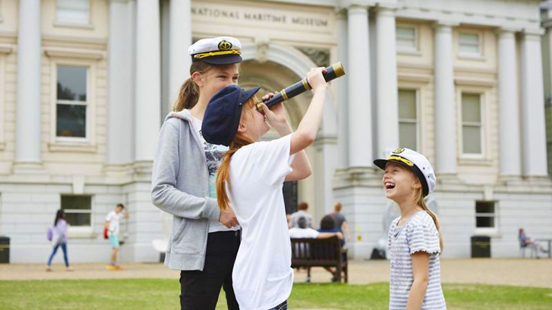 Enjoy plenty of hands on activities for children of all ages at the National Maritime Museum. © National Maritime Museum, Greenwich, London, 2026. Image courtesy of Visit Greenwich. A photo of three children of different ages laughing together outside the National Maritime Museum, each wears a sailor cap and one is looking through a telescope