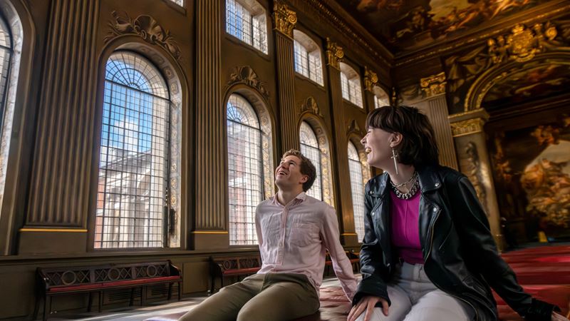 The Painted Hall inside the Old Royal Naval College in Greenwich is a must-visit. Credit: London & Partners/Michael Barrow. Image courtesy of London & Partners. A photo of a man and woman sat down inside the Painted Hall smiling together whilst admiring the painted walls and ceiling