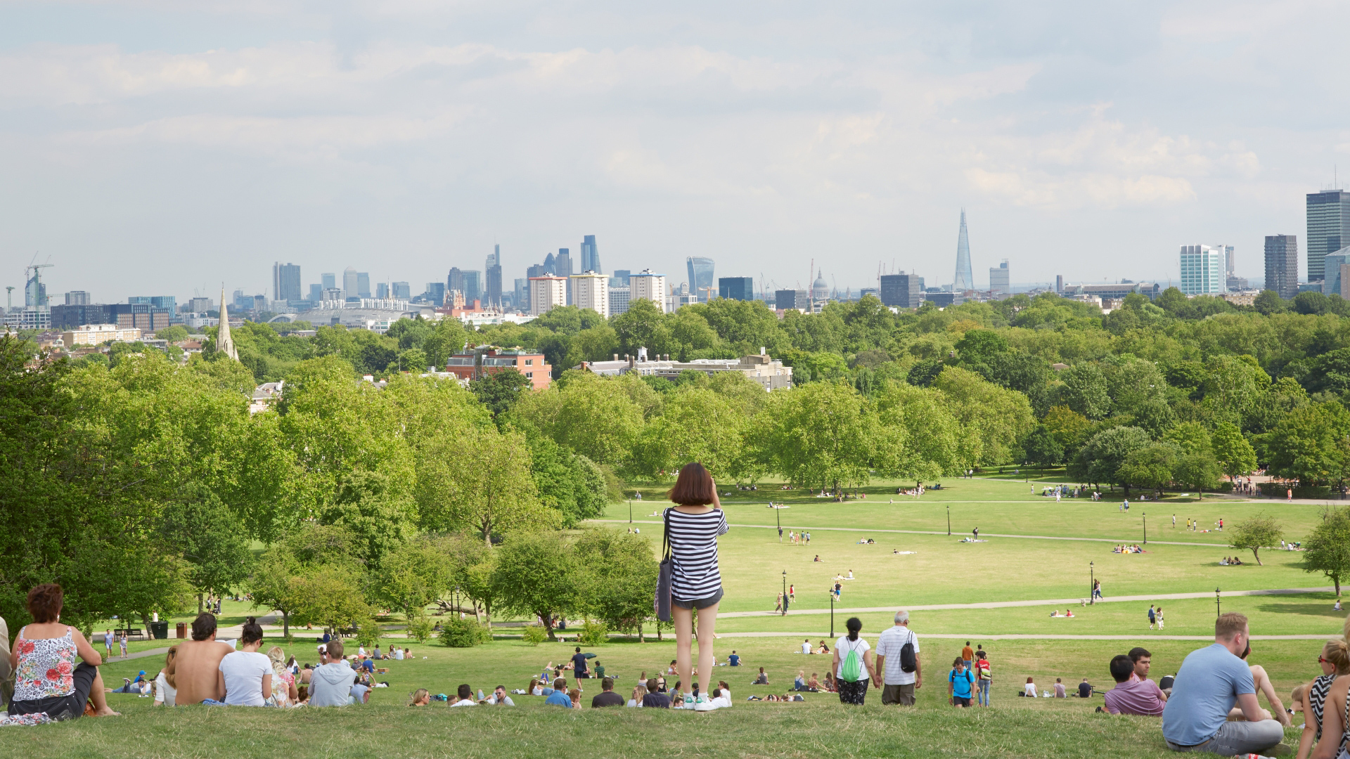 Snap a photo of one of the best London skyline views at Primrose Hill. Credit: Shutterstock. Image courtesy of Shutterstock. A photo of lots of people sat on Primrose Hill facing the London skyline on a sunny summers day with one woman stood up taking a photo