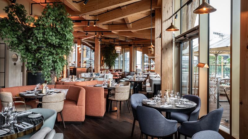 Tuck into classic signature dishes from celebrity chef Gordon Ramsay in Stratford. Credit: Bread Street Kitchen. Image courtesy of Bread Street Kitchen. Interior dining room at Bread Street Kitchen in Stratford, London, featuring floor-to-ceiling windows, orange and grey seating, industrial-style lighting, wooden ceiling and hanging plants.