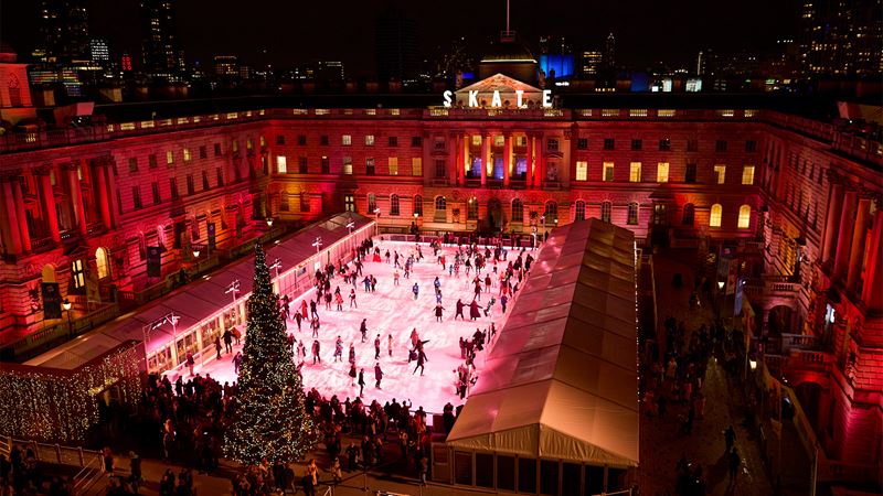 Skate at Somerset House. Image courtesy of Owen Harvey/Somerset House. Skaters glide across the ice at Somerset House against a glittering spruce tree and red lights
