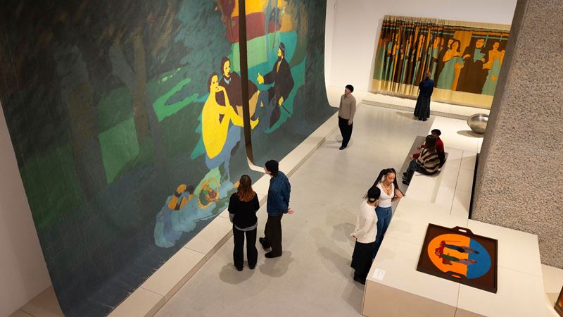 Installation view, Beatriz González at the Barbican, London. Image credit - Barbican Art Gallery, David Parry © Beatriz González. Multiple people wandering around exhibition space with colourful artworks at Barbican Art Gallery.