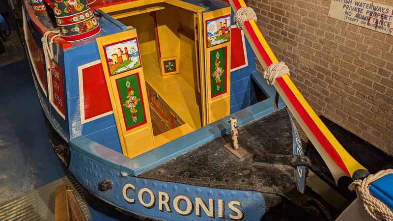 Learn about the history of canals and life aboard canal boats, such as the Coronis, at the London Canal Museum. A coloufully painted canal boat in blue red, yellow and green, with the name "Coronis" painted on the side.