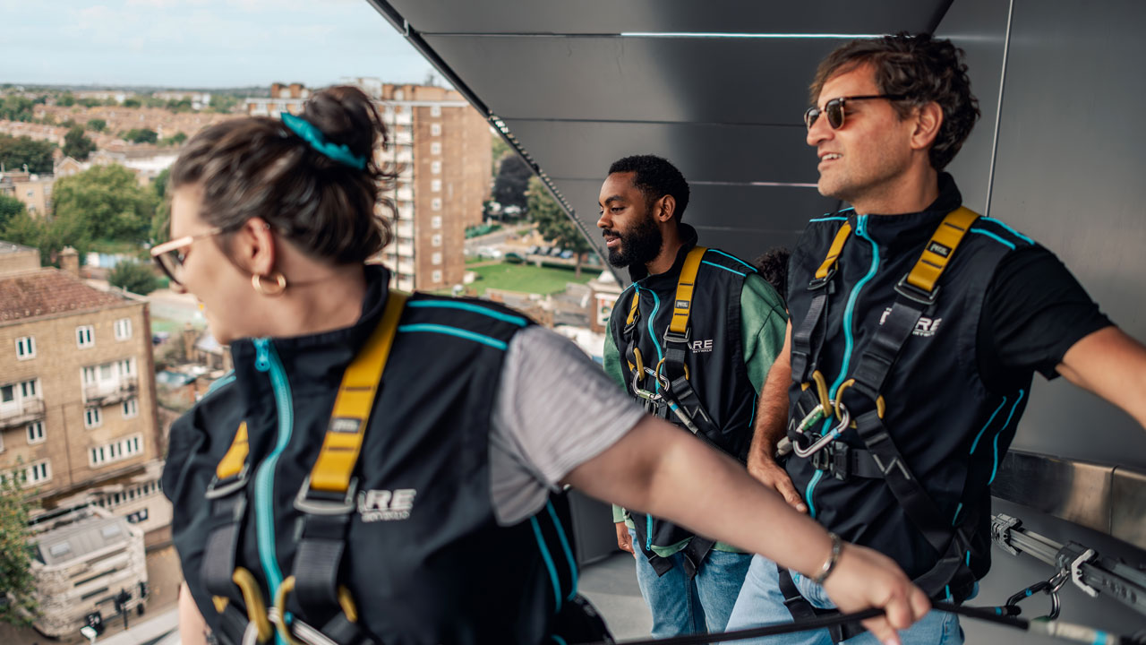 Three people look down at the landscape while holding a safety rope on their way to the top of Tottenham Hotspur Stadium.