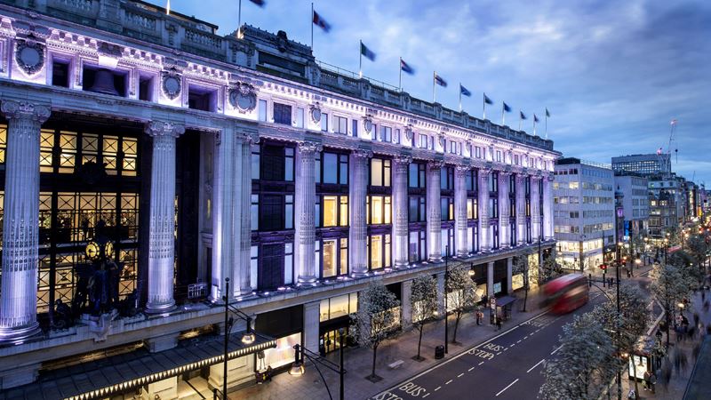 Explore Selfridges, one of London’s most iconic department stores, celebrated for fashion, food and culture © Andrew Meredith/Selfridges. Image courtesy of Selfridges. A panoramic view from high of the facade of Selfridges, lit up in purpley-white lights, with Oxford Street below.
