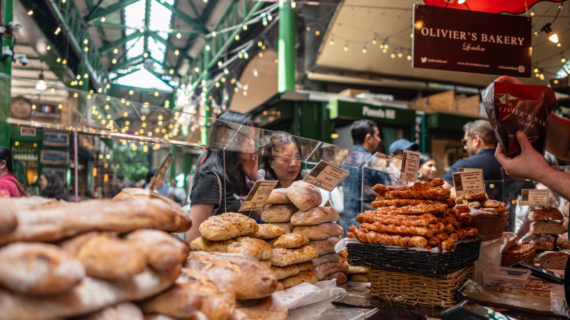 Explore stalls filled with foodie delights at Borough Market. © Michael Barrow/London & Partners. People being served at a bakery stall full of bread and baked goods within Borough Market, with the famous green struts of the market in the background.