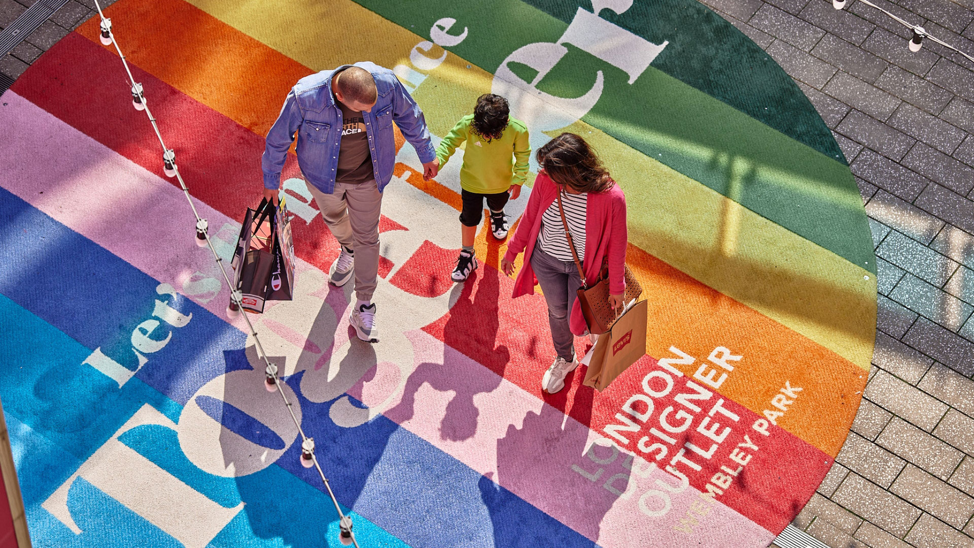Bring the whole family to London Designer Outlet for a day of shopping and more fun activities on offer in the area. Credit: London Designer Outlet. Image courtesy of London Designer Outlet. A bird's-eye photo of a couple holding hands as they walk with their young child on top of a giant rainbow-coloured London Designer Outlet floor sticker with shopping bags in their hands