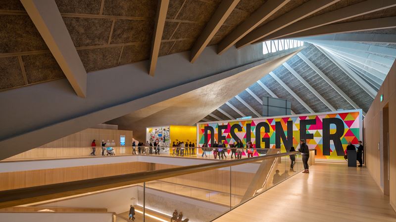Discover the designs that have helped shape our cultural landscape at the Design Museum. Credit: Jon Reid. Image courtesy of visitlondon.com. A large billboard with colourful triangles and the word "Designer" emblazoned across it, on the top floor of the Design Museum, with people walking past it.