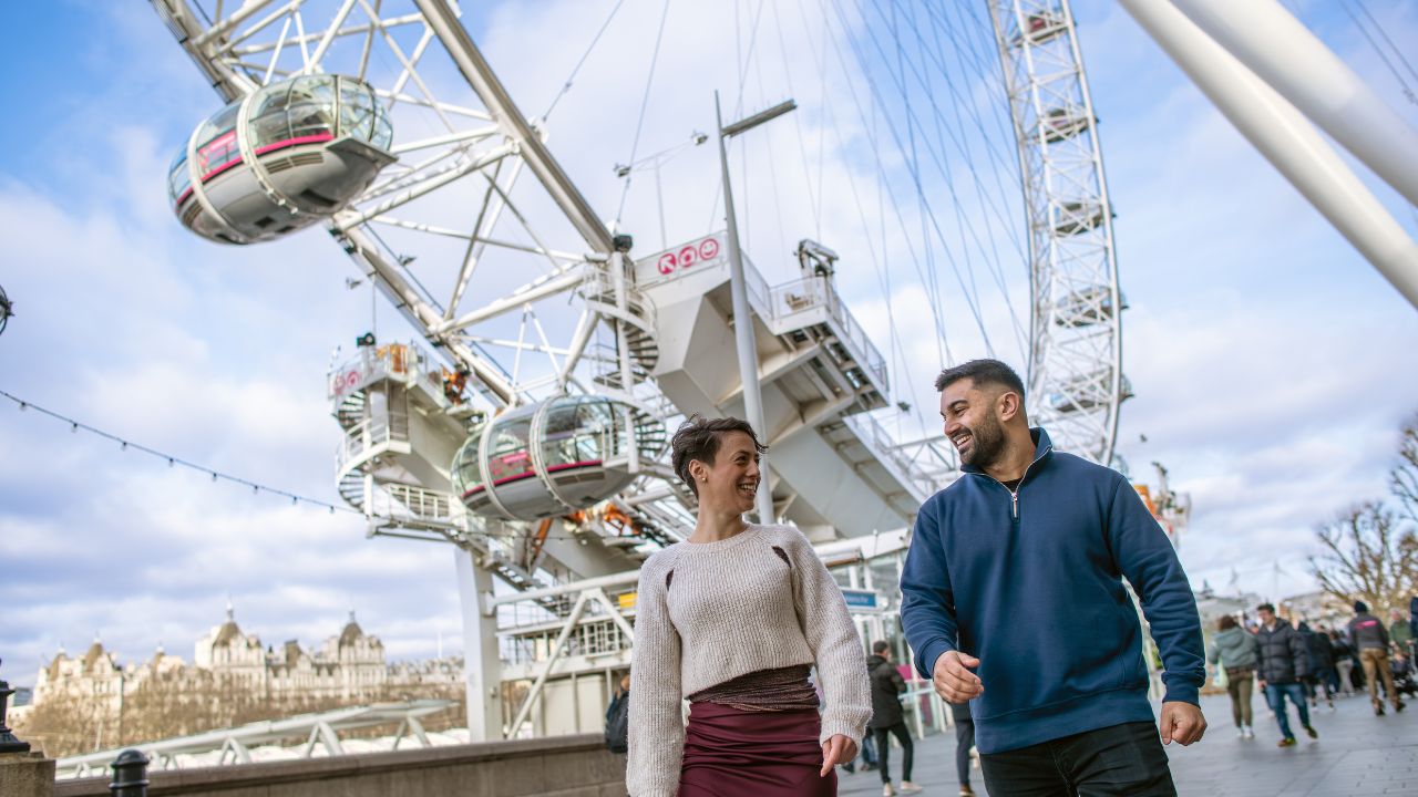 Enjoy 360-degree views of London with a trip on the London Eye. © London & Partners/Michael Barrow. Two people walk in front of the pods of the London Eye.