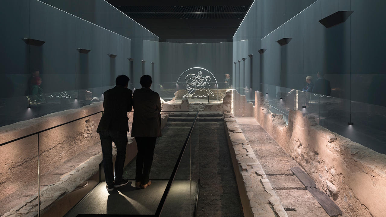Step back into Roman times at the London Mithraeum. Image courtesy of London Mithraeum. Two people look over the ruins of the Temple of Mithras, at the dimly lit London Mithraeum.