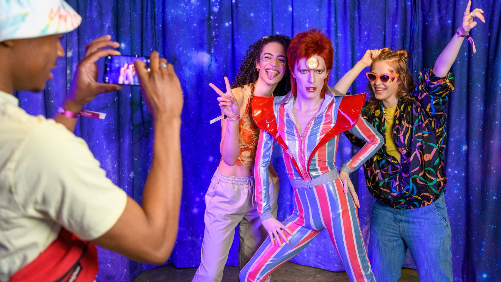 Strike a pose with pop icon David Bowie at Madame Tussauds London. Image courtesy of Madame Tussauds London. wax figure model of David Bowie, with his iconic red hair and blue and red stripey suit. a man is taking a photo of two women posing next to the model.