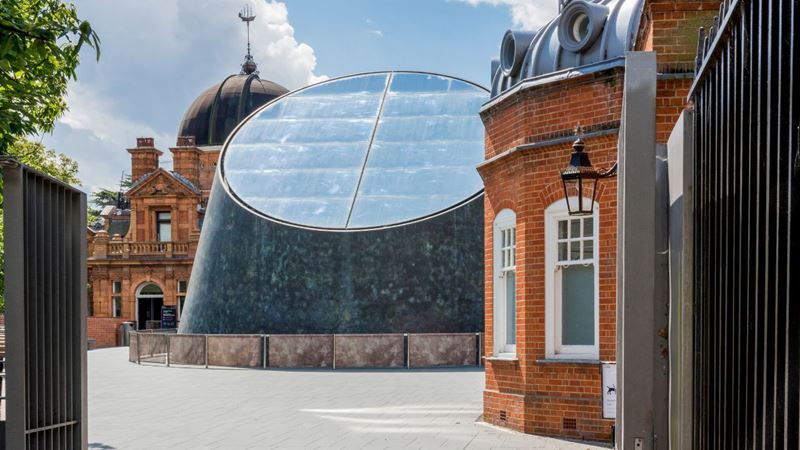 See our world and beyond from a new perspective at the Peter Harrison Planetarium. Image courtesy of Royal Museums Greenwich. Large grey and glass building in greenwich against two red brick buildings on the site of the peter harrison planetarium.