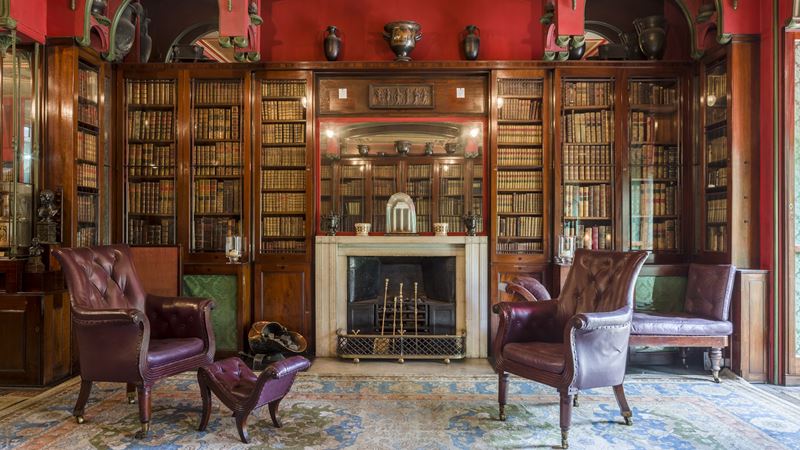 The fireplace in the Library Room at Sir John Soane's Museum. Credit: Gareth Gardner. Image courtesy of Sir John Soane's Museum. Cosy library room packed with book shelves, a fireplace, leather armchairs, red walls and a big colourful carpet.