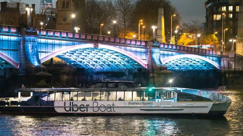 See London lit up on a river Thames cruise. Image courtesy of Golden Tours. A black and white uber cruise boat sails underneath a London bridge lit up with blue lights